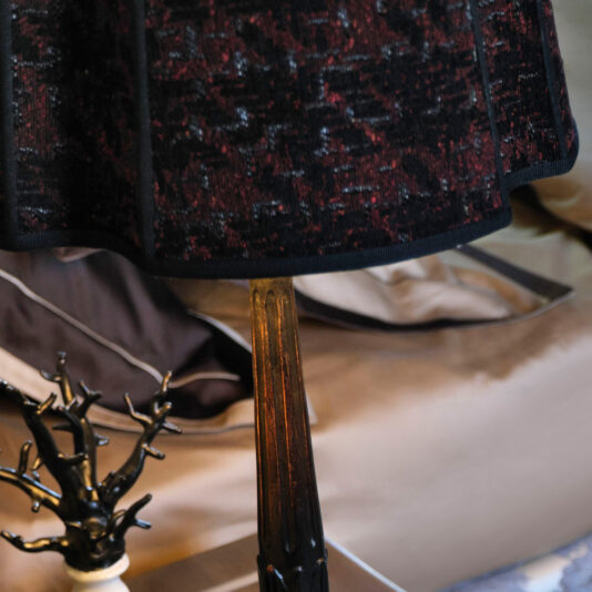 Large Luxury Carved Table Lamp Close-up of a Large Luxury Carved Table Lamp featuring a dark red and black speckled lampshade. The metallic base is placed on a table next to a decorative black tree-shaped object. In the background, there are pillows and a neatly made bed.