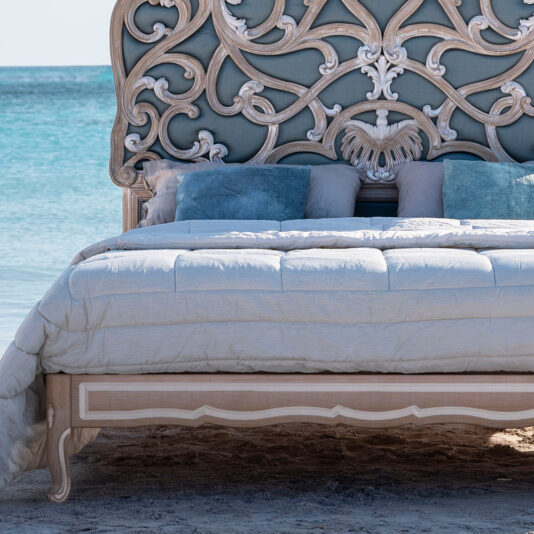 Ornate Rococo Style Bed With Tall Headboard The Ornate Rococo Style Bed with Tall Headboard sits on a beach with turquoise waters in the background. The bedding is in light shades, and several blue and gray decorative pillows are arranged against the headboard. The bed frame features elegant carvings.