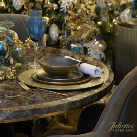 A luxurious dining room holiday table is set with gold-rimmed plates, a dark bowl, a rolled white napkin, and blue glassware on a dark marble surface. The background features a decorated Christmas tree with gold, silver, and blue ornaments. "Juliettes Interiors" logo is visible.