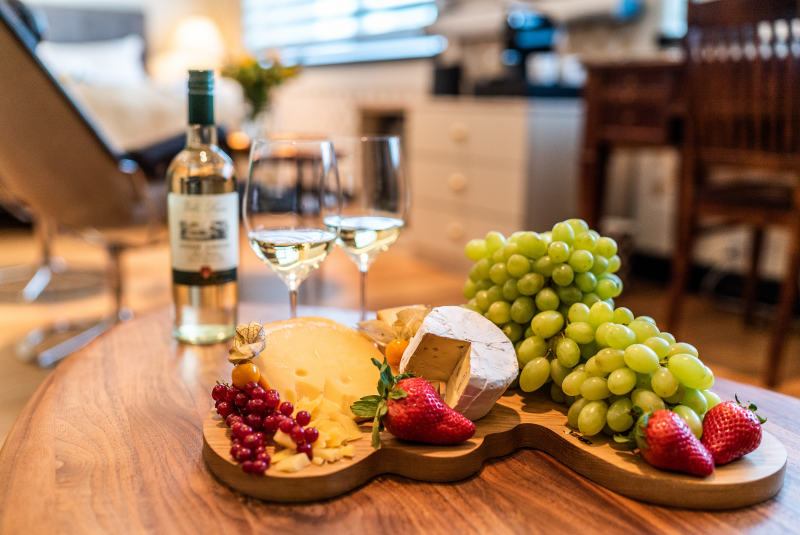 A cozy indoor setting featuring a wooden table with a wine bottle, two filled wine glasses, a cheese platter with assorted cheeses, red currants, strawberries, green grapes, and a bed with a reading chair in the background – truly the perfect scene for any luxury giveaway.