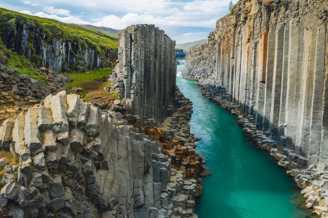 A stunning canyon featuring towering basalt columns on either side, with a turquoise river flowing between them. The landscape is lush with greenery on the left side, while the right side showcases the vertical rock formations under a partly cloudy sky—a true luxury giveaway from nature.