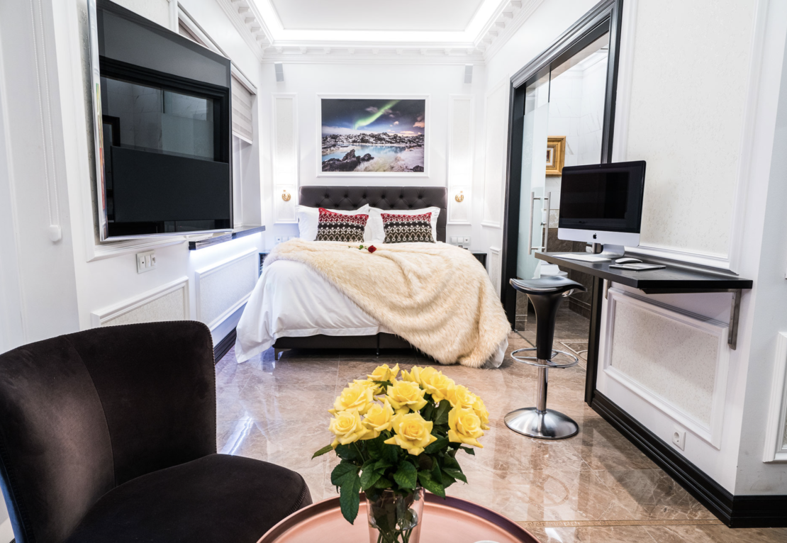 A modern bedroom with a queen-size bed, decorated with a fluffy beige blanket and patterned pillows, radiates an understated elegance. A black armchair and a round glass table with yellow roses sit in the foreground. To the right, there's a wall-mounted computer desk and chair, perfect for our luxury giveaway setup.