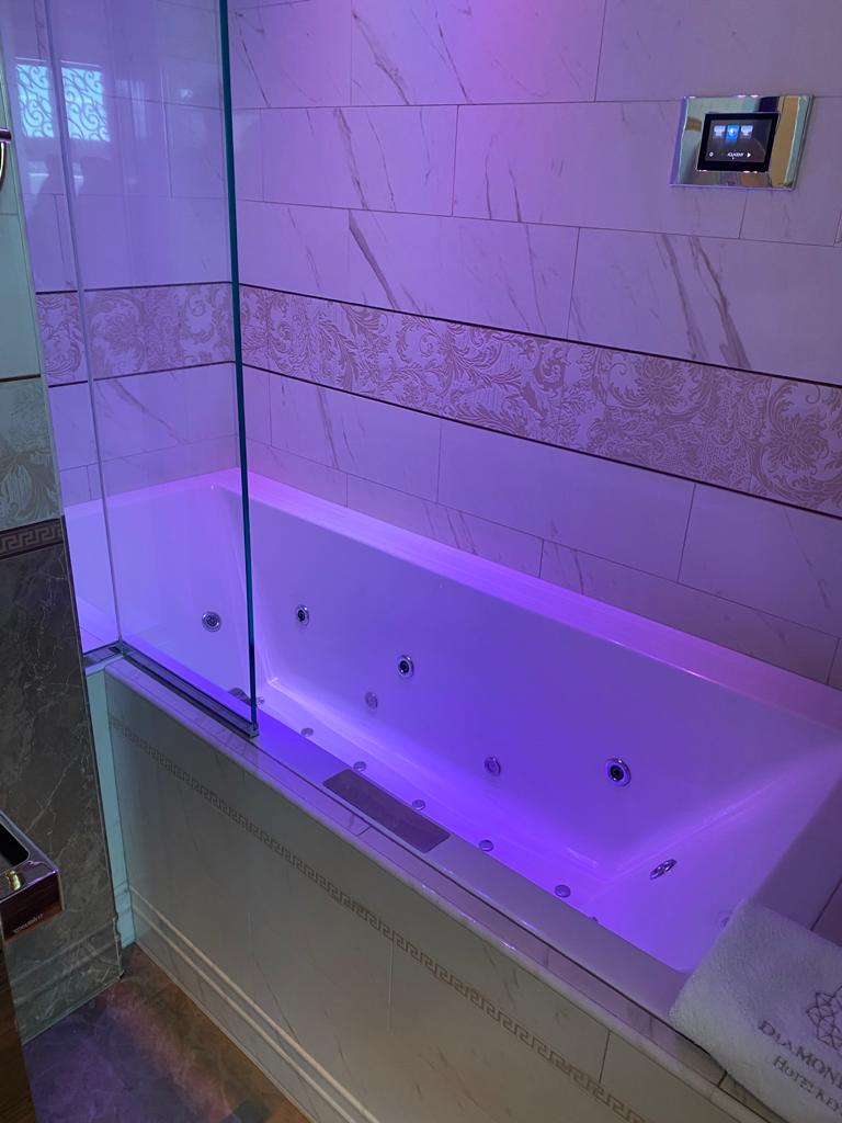 A modern white bathtub with jacuzzi jets is installed in a bathroom with marble walls and a glass screen. The bathtub is illuminated with purple LED lights, exuding a sense of luxury. As part of our exclusive luxury giveaway, a folded towel with a floral design adorns the bathtub edge.