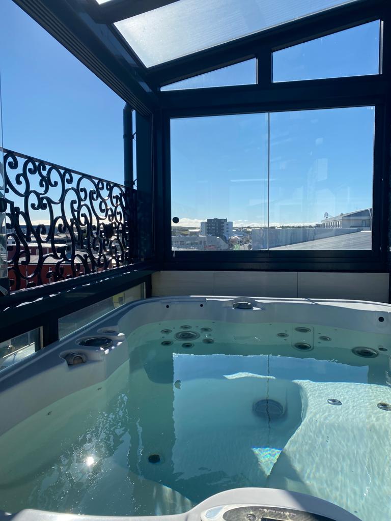 A luxurious outdoor hot tub with clear water is enclosed within a glass balcony. Decorative black railings are visible on the left, and a sunny cityscape can be seen through the glass windows towering over buildings in the background, offering a perfect setting for our exclusive luxury giveaway experience.