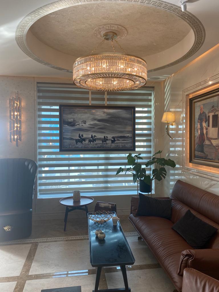 A luxurious living room with a crystal chandelier, leather couch, and modern coffee table. A painting of horse riders hangs on the wall, beneath which is a large window with zebra blinds. A potted plant and a unique wall light also decorate the space—an ideal setting for our exclusive luxury giveaway.