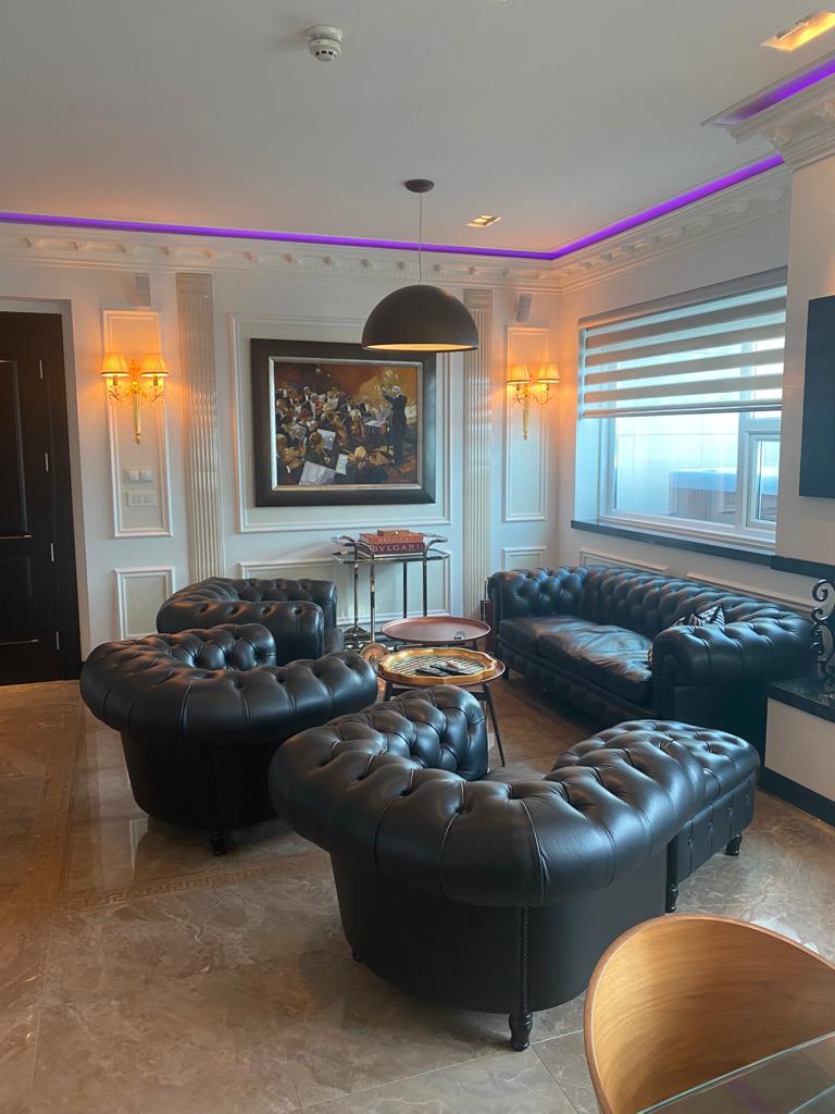 Luxurious living room featuring black leather chesterfield sofas and armchairs, a central coffee table with a chessboard, a large framed painting on the wall, elegant wall lighting, marble flooring, and a window with striped blinds—perfect for any luxury giveaway setting.