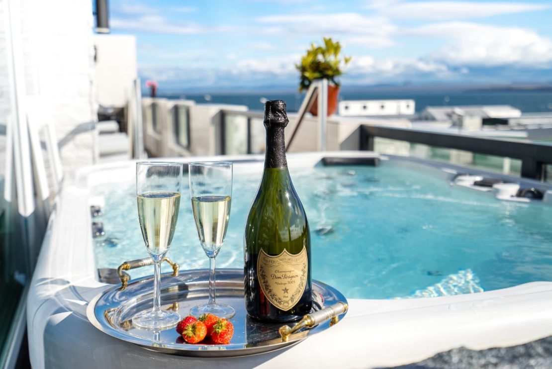 A tray with a bottle of champagne, two filled flutes, and strawberries is placed on the edge of a hot tub. The hot tub is on a balcony overlooking a scenic view of the ocean and distant mountains under a blue sky—an idyllic scene for our luxury giveaway.