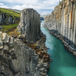 A stunning landscape featuring the Studlagil Canyon in Iceland, showcasing towering basalt columns on either side of a serene, turquoise river. The dramatic rock formations create a narrow passage, with grassy hills and a partly cloudy sky in the background—a true masterpiece worthy of a luxury giveaway.