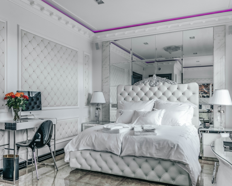 A luxurious bedroom features a white tufted bed with decorative pillows and folded towels. The room includes mirrored walls, elegant lighting, a large bouquet of red and orange roses on a desk, and a computer. The ceiling has a purple light accent—a perfect setting for our luxury giveaway.