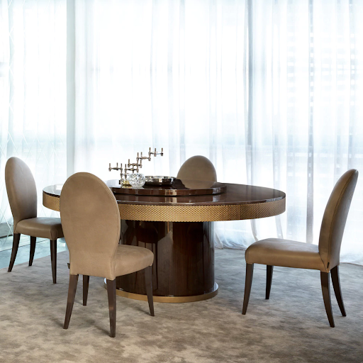 A round dining table with a wooden top and brass trim is set against a large window with sheer white curtains, seamlessly fitting into various home colour schemes. Four beige upholstered chairs surround the table. On the table is a decorative candelabrum and glass bowl. The floor features a light gray carpet.