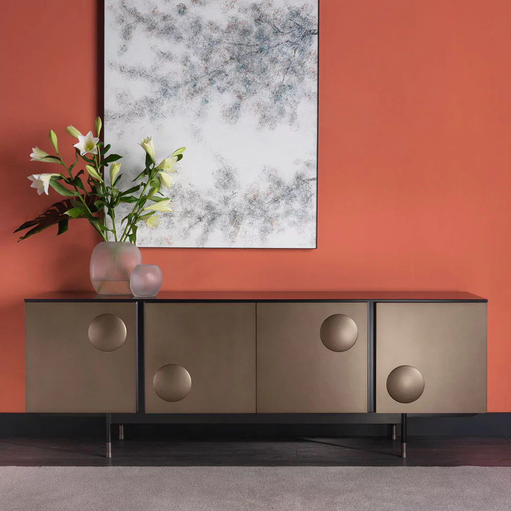 A modern sideboard with a bronze finish stands against an orange wall, seamlessly integrating into diverse home colour schemes. On top of the sideboard are a glass vase with white lilies and a frosted vase. Above the sideboard hangs an abstract painting dominated by shades of white and gray.