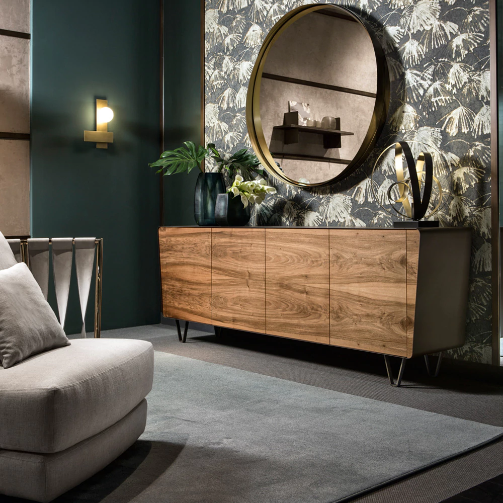 A modern living room featuring a wooden sideboard beneath a large round mirror with a gold frame exemplifies refined home colour schemes. The wall boasts leafy patterned wallpaper, while various vases and a decorative sculpture adorn the sideboard. A cushioned armchair and wall sconce complete the scene.
