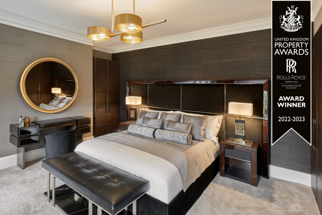 A luxurious bedroom featuring a large bed with elegant grey bedding, a black upholstered headboard, and modern bedside tables with lamps. There's a circular mirror above a stylish desk and chair. A "United Kingdom International Property Award 2022-2023" banner is displayed in the corner.