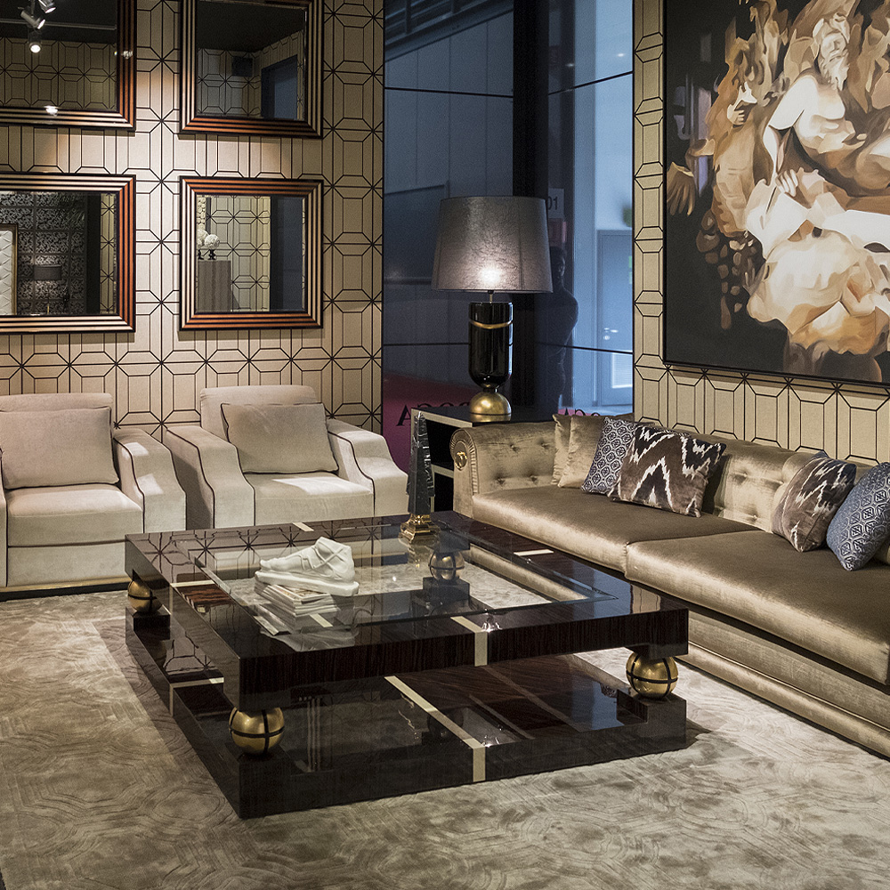 A modern, stylish living room with beige geometric wallpaper and a plush rug. It features a beige sectional sofa, two matching armchairs, a black glossy coffee table, multiple framed mirrors on the wall to maximize lighting tips, a black and gold lamp, and a large abstract artwork.