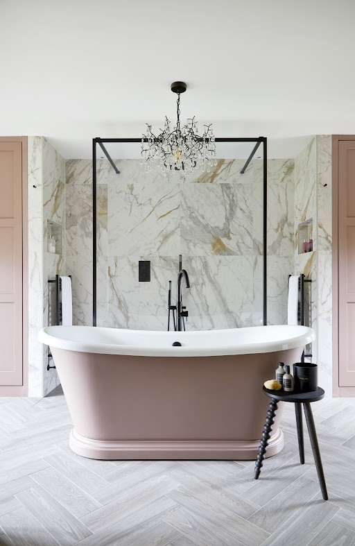 This image showcases a luxurious bathroom with a freestanding pink bathtub and a chandelier above. The back wall, adorned with marble tiles, perfectly complements the room's sophisticated home colour schemes. A herringbone-patterned floor adds texture, while a black side table beside the tub holds toiletries.