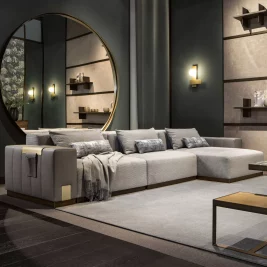 A contemporary living room featuring a large gray sectional sofa with throw pillows and a blanket draped over the arm. A round mirror on the wall reflects the room's cohesive home colour scheme, which includes dark wood flooring, built-in shelves with decorative items, and wall-mounted lights.