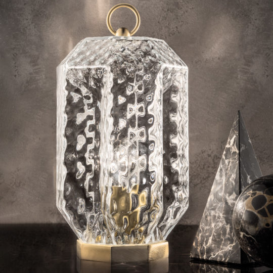 The Lantern Style Murano Glass Lamp, featuring a hexagonal glass design with a brass base and handle, is placed on a reflective dark surface alongside a black marble pyramid and sphere. The background is set against a grey textured wall.