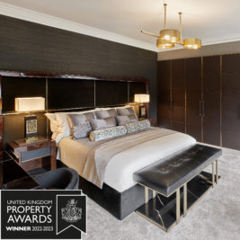 A luxurious contemporary bedroom features a large bed with plush bedding and multiple throw pillows. A stylish bench sits at the foot of the bed. Modern bedside tables, lamps, and a wardrobe complete the look. An award graphic reads "International Property Award Winner 2022-2023" in the bottom left corner.