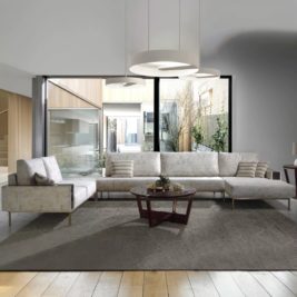 A modern living room with a long L-shaped white sofa, three round white ceiling lights, and a round wooden coffee table on a gray rug. Large windows offer a view of an outdoor area comprising greenery and a contemporary building, perfectly embodying 2023 interior design trends.