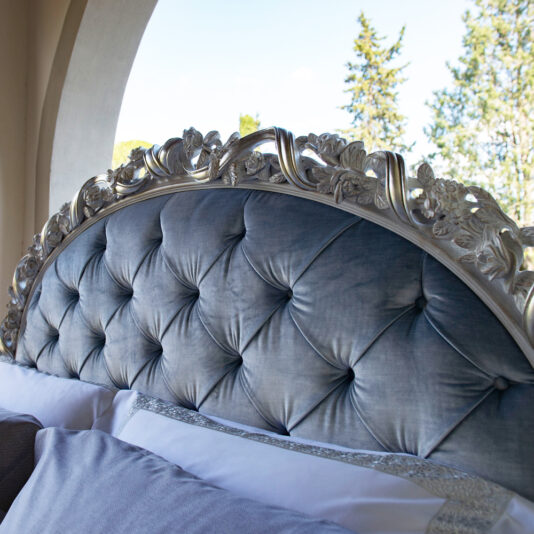 Ornate Carved Button Upholstered Bed A close-up of the Ornate Carved Button Upholstered Bed, featuring intricate silver floral carvings and blue button-tufted upholstery. The bed is adorned with plush pillows and sits near a large window that provides a view of trees and a bright sky.
