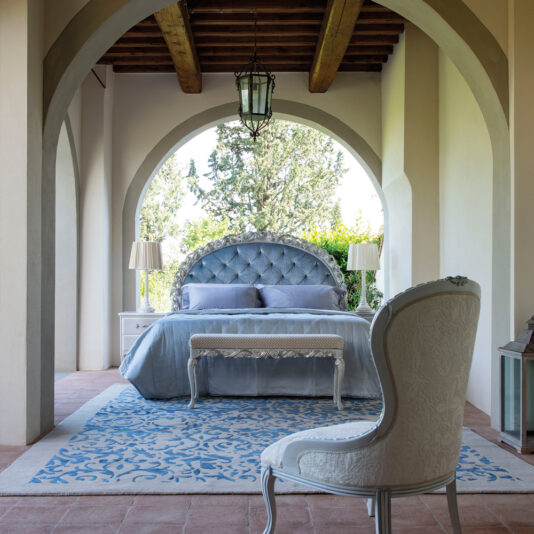 Ornate Carved Button Upholstered Bed An elegant bedroom with a high, arched ceiling features an Ornate Carved Button Upholstered Bed in luxurious gray, complemented by a matching comforter set and two white bedside tables with lamps. A white ornate chair faces the bed on a blue and white patterned rug, while an ornate lantern hangs from the ceiling.