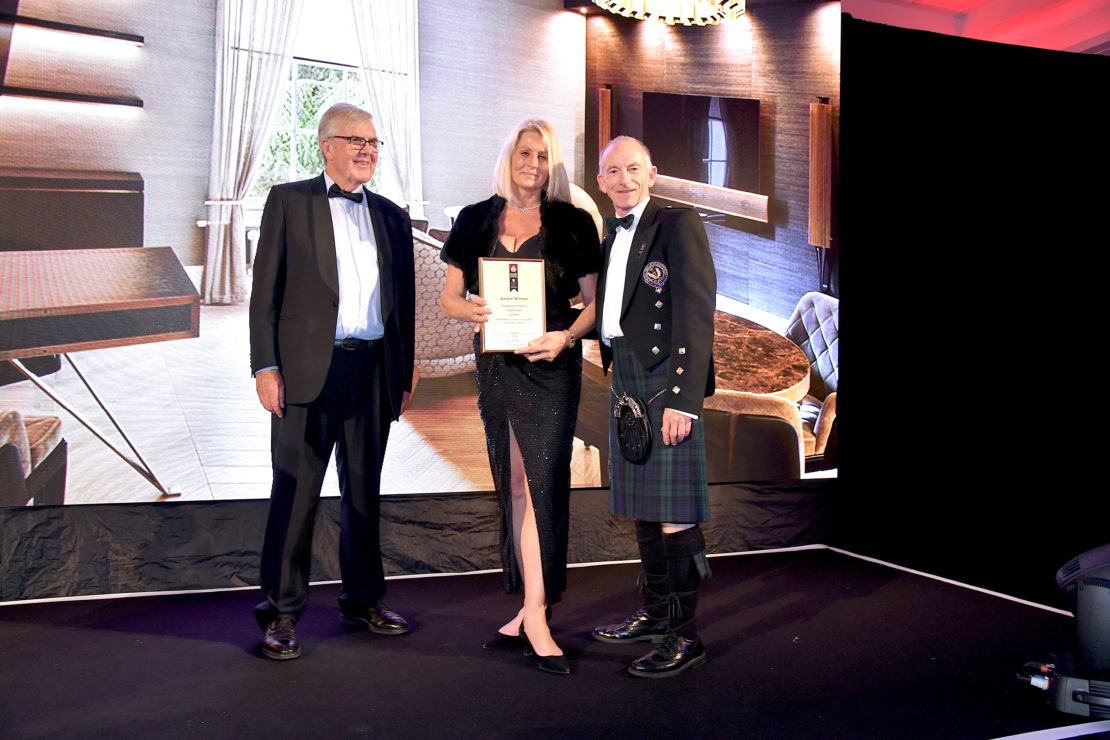 Three people are standing on a stage, with a woman in the center holding a framed international property award. The man on the left is wearing a suit and bow tie, while the man on the right is dressed in a traditional Scottish kilt. A large screen displays a modern room behind them.