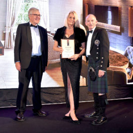 Three people pose for a photo at an event. The woman in the center holds a framed certificate for an international property award and wears a black evening gown. The men on either side of her, one in a suit and the other in a kilt and jacket, smile for the camera.