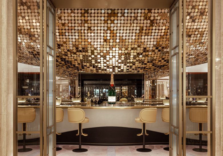 Elegant, modern bar interior with a luxurious gold-toned ceiling made of circular discs, high stools, a central counter with a large bottle of champagne, and glass doors framing the entrance, conveying a sophisticated ambiance.