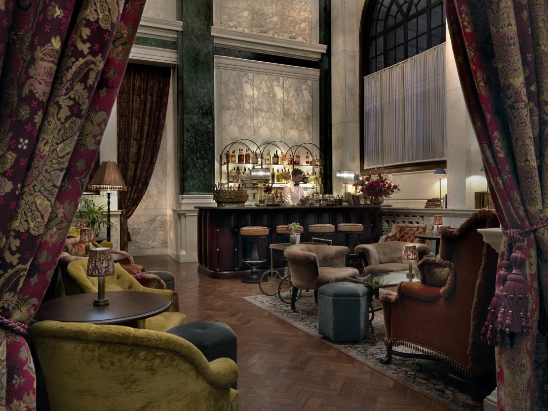 A cozy, upscale hotel lounge with ornate curtains pulled to the side, revealing a warmly lit bar area. The room features elegant furniture including velvet armchairs, a couple of small round tables, a floor lamp, and rich dark wood paneling. A bartender is seen behind the bar.