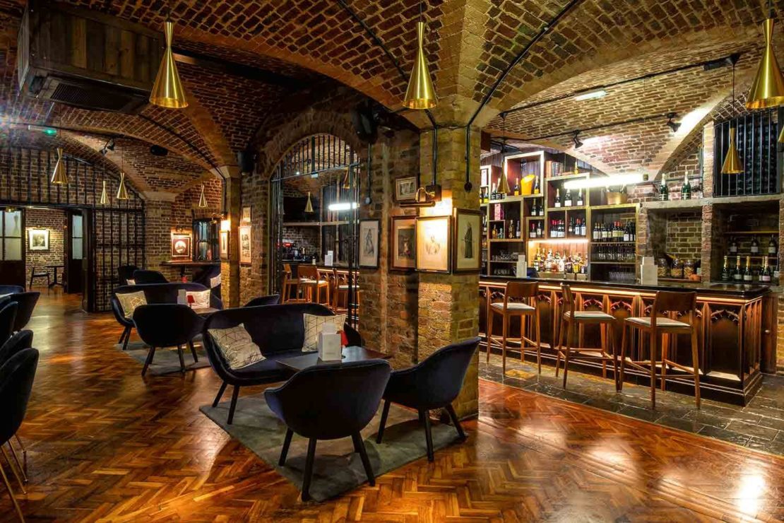 A cozy, dimly-lit bar with exposed brick walls and archways. The space includes a counter with barstools, several round tables with black chairs, and plush seating. Elegant pendant lights hang from the ceiling, and shelves with bottles of alcohol line the back wall.