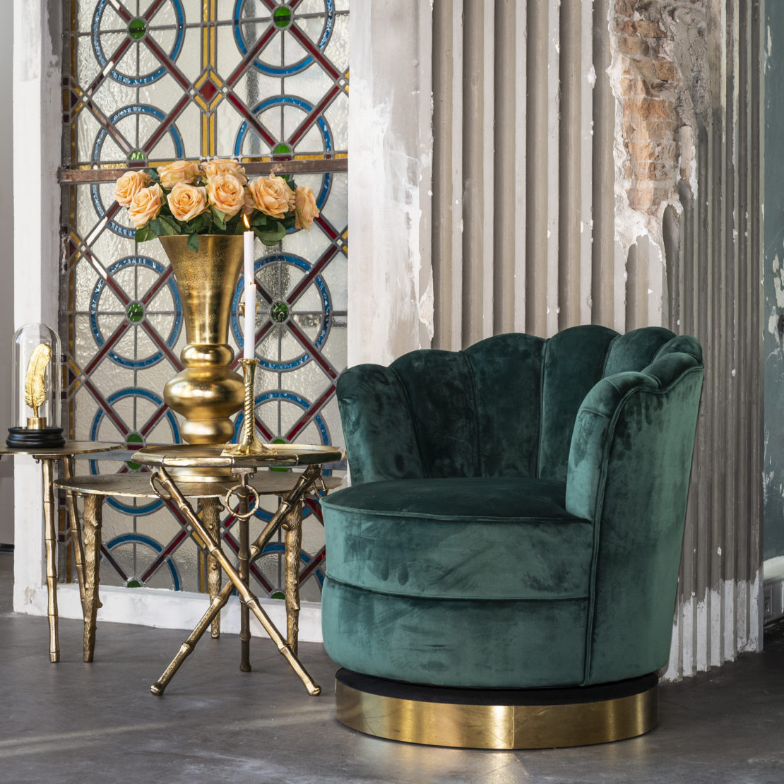 An elegant interior scene featuring a plush green velvet chair with a scalloped back, a round glass-top table with gold accents holding an ornate vase filled with peach roses, and a geometric stained glass window in the background. The walls are weathered and textured.