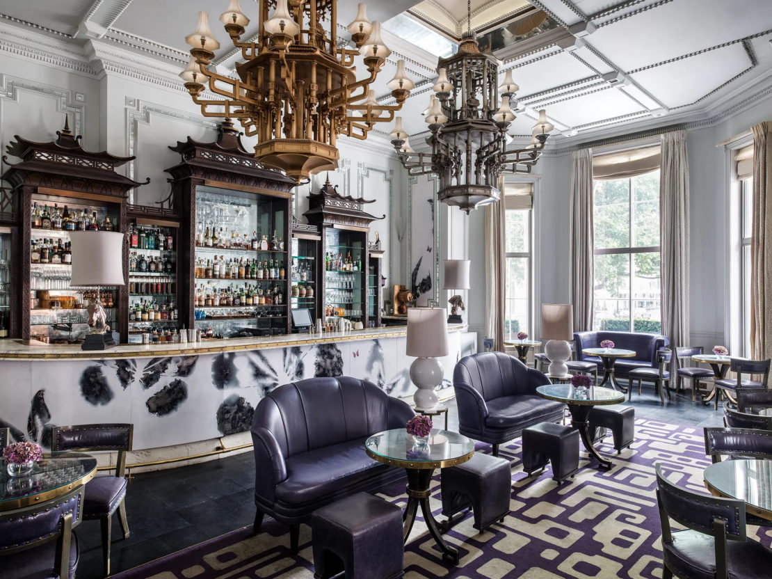 A luxurious bar lounge with an elegant and modern design. The room features a marble bar center, dark leather seating, small round tables, ornate chandeliers, and large windows that let in natural light. The decor blends classic and contemporary elements seamlessly.