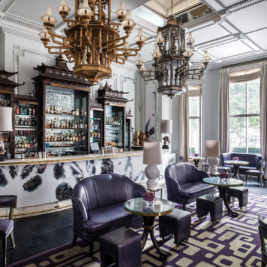 A luxurious bar lounge with an elegant and modern design. The room features a marble bar center, dark leather seating, small round tables, ornate chandeliers, and large windows that let in natural light. The decor blends classic and contemporary elements seamlessly.