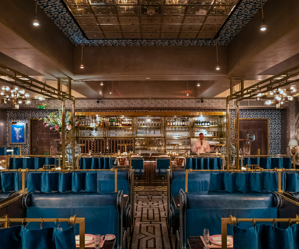 An elegant restaurant interior with plush blue booth seating, gold accents, and intricate lighting fixtures. The bar area features an ornate metallic backsplash and a variety of glassware. The ceiling has a reflective, patterned metallic design.