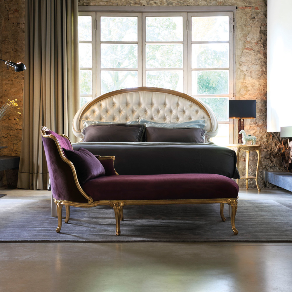 A stylish bedroom features a luxurious bed with an ornate tufted headboard and dark bedding. In front, there's a purple velvet chaise lounge with gold accents. Large windows and a stone accent wall add a warm, rustic touch to the room, while hints of viva magenta bring an unexpected pop of color.