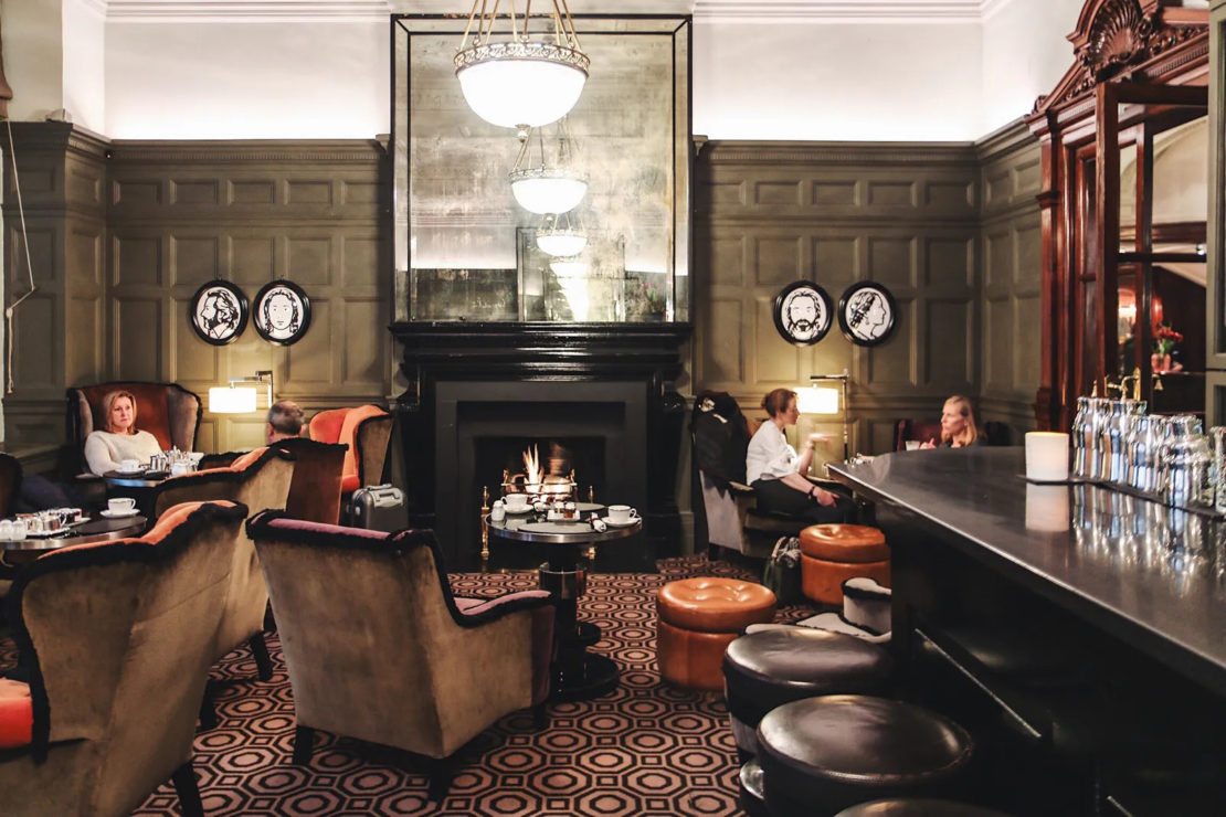 A cozy, elegant lounge with plush armchairs, a lit fireplace, and a large mirror above. People are seated, engaged in conversation and enjoying beverages. The room is warmly lit with chandeliers and has a vintage feel with dark wood paneling and decorative plates.