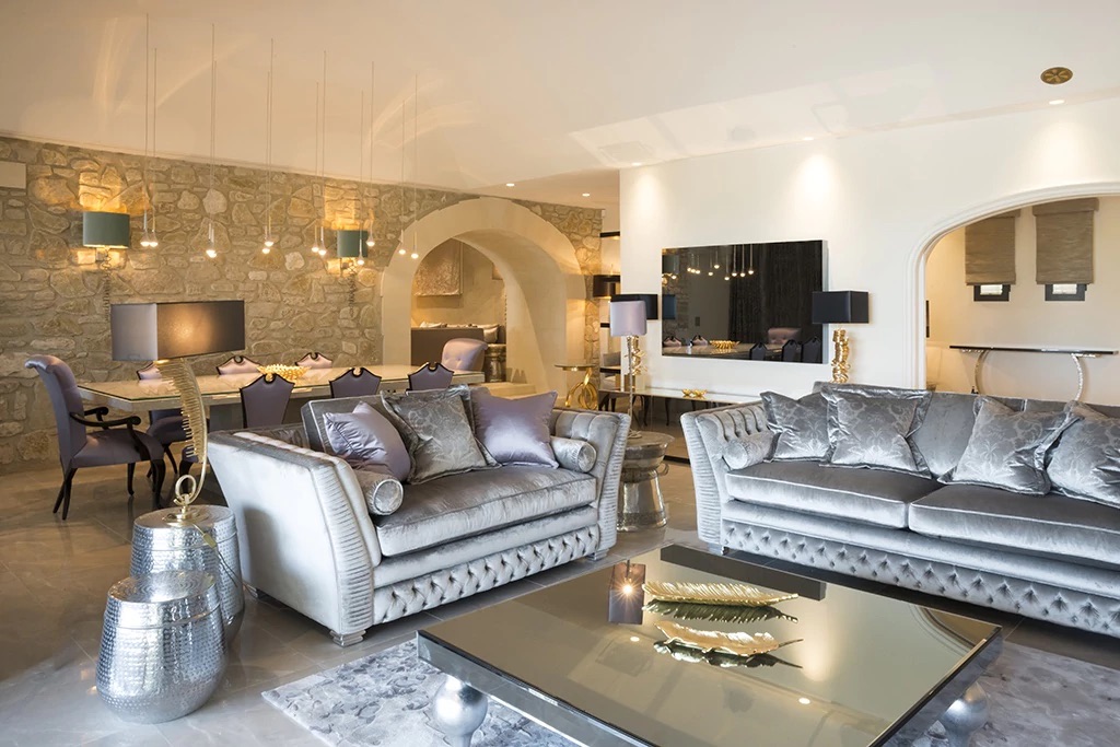 A luxurious living room with plush silver sofas and a mirrored glass coffee table. The space features a stone accent wall, modern lighting fixtures, an elegant dining area with upholstered chairs in digital lavender, and decorative elements like metallic vases and a golden feather centerpiece.