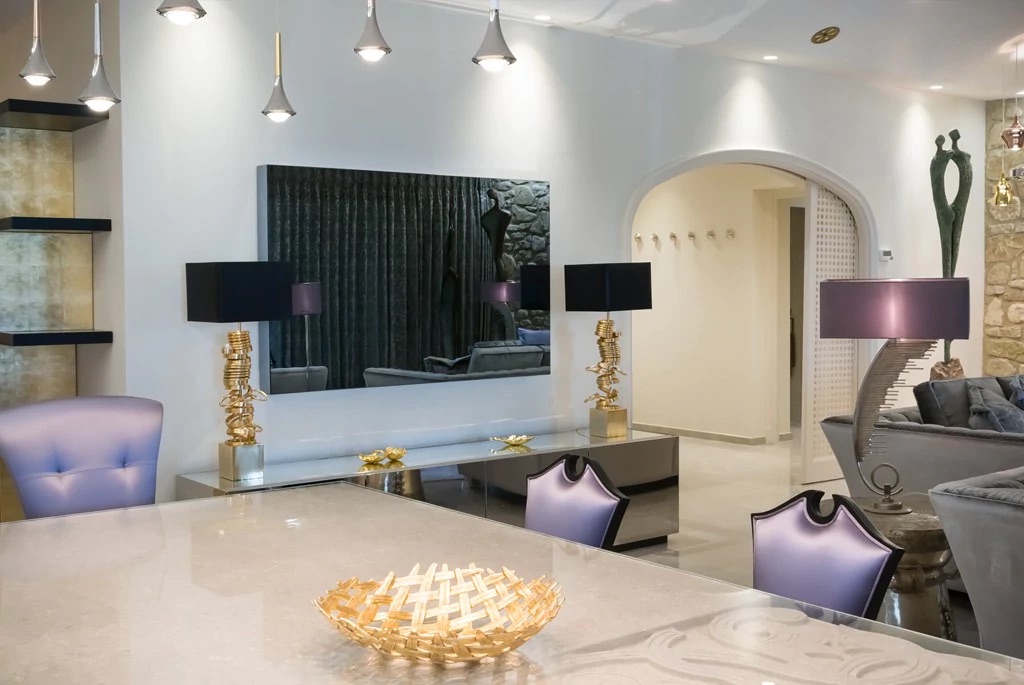 A modern, stylish living room with a mix of contemporary and classic elements. The room features elegant furniture, including grey sofas, digital lavender metallic side tables, and unique, artistic table lamps. A large wall mirror and arched doorway add sophistication to the space.