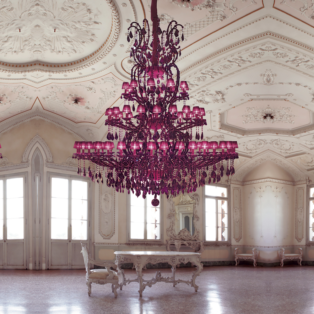 An opulent room features an intricately designed, large purple chandelier hanging from a richly decorated ceiling. The space includes elegant white furniture and large windows, allowing natural light to pour in, highlighting the room's ornate architectural details touched with hints of viva magenta.