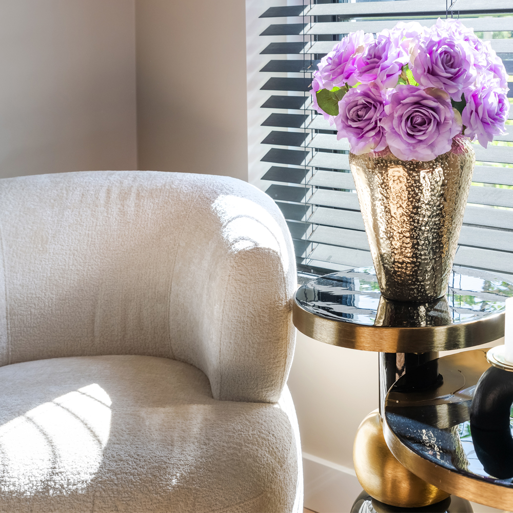 A cozy living space featuring a plush beige armchair beside a round, tiered side table. On the table rests a gold textured vase with vibrant purple roses. A window with dark horizontal blinds casts shadows tinged with digital lavender hues across the room.