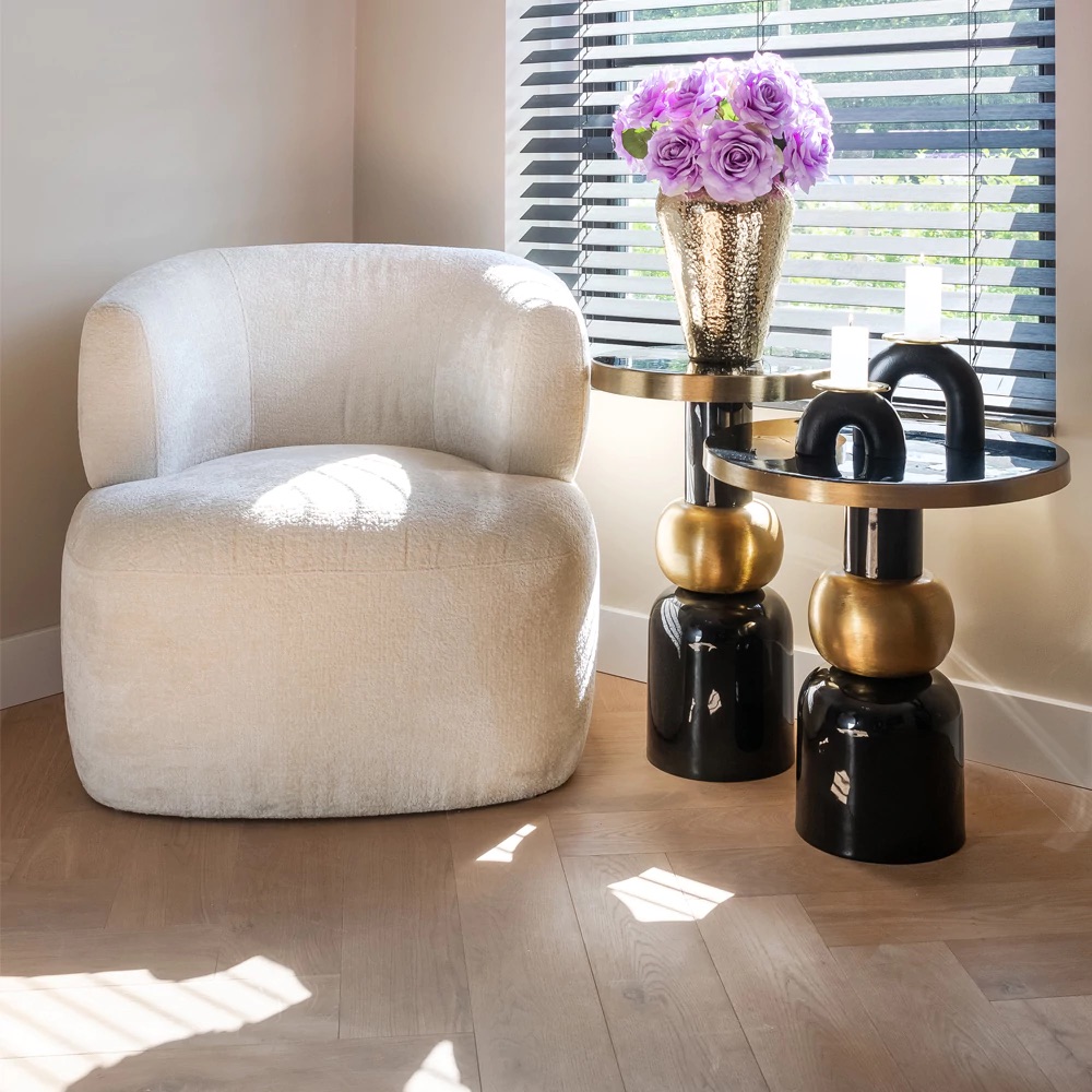 A cozy white armchair sits beside two modern black and gold side tables. One table holds a vase of vibrant digital lavender flowers. Sunlight filters through the blinds, casting shadows on the light wooden floor. The scene conveys an inviting and stylish corner for relaxation.