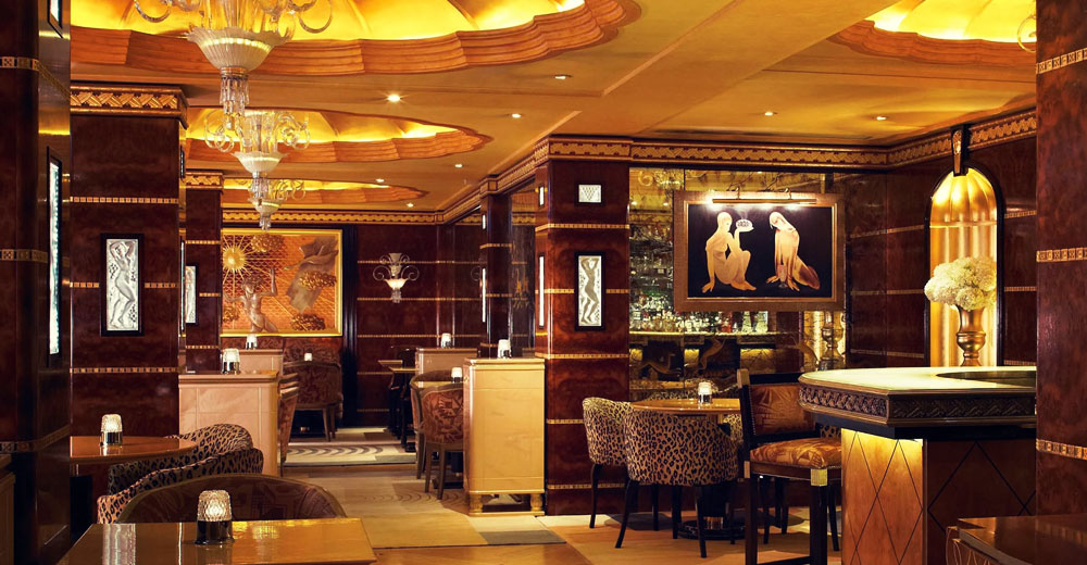 A sophisticated, warmly lit lounge with ornate ceiling designs, plush seating, and decorative wall art. The lounge features marble-top tables, patterned armchairs, and ambient lighting creating an elegant and inviting atmosphere.