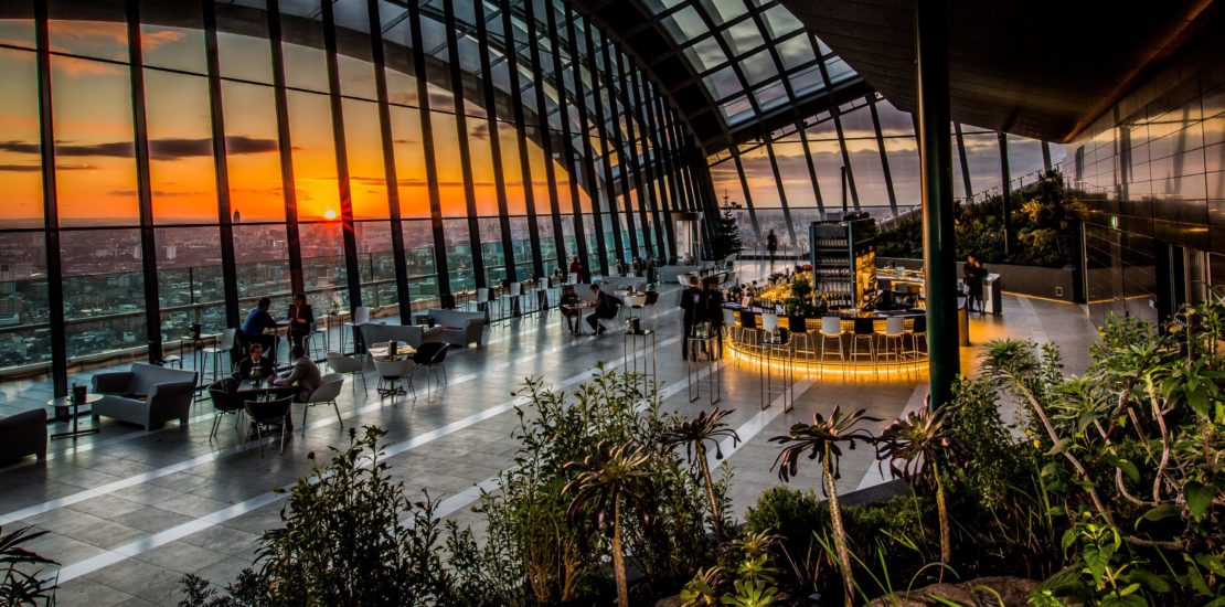 A modern rooftop bar with large arched windows offering a panoramic view of the city at sunset. Patrons seated at tables and the bar, surrounded by indoor plants, enjoy the vibrant atmosphere. The sky lights up with orange and purple hues as the sun sets.