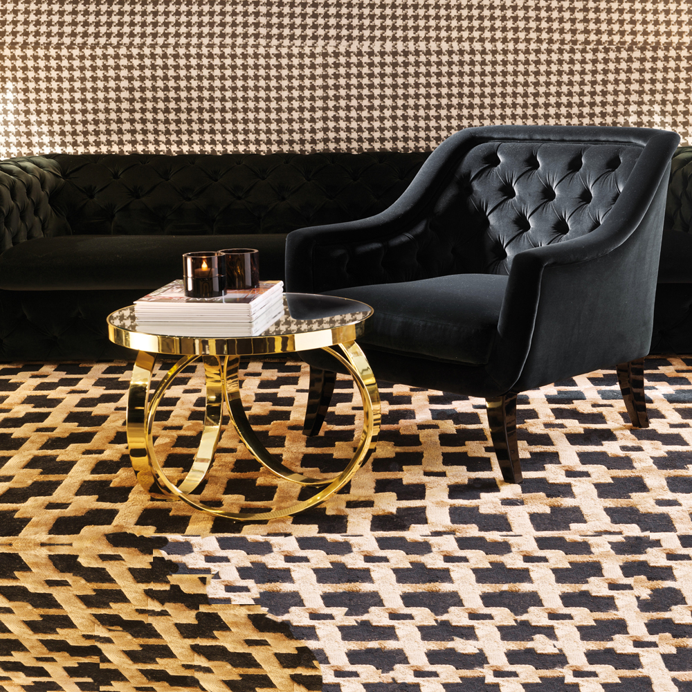 A stylish living room with a black tufted armchair and matching sofa. A small golden accent table with a circular glass top holds two candles and a stack of books. The room, adorned with houndstooth patterned wallpaper and rug in black and beige tones, exudes an elegant black and gold theme.