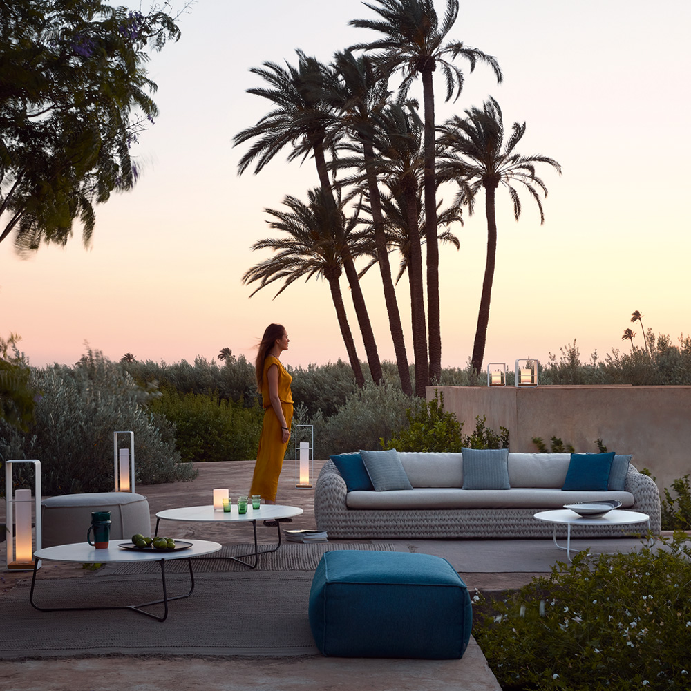 A person in a yellow outfit stands near several palm trees in an outdoor lounge area at sunset. The luxury exterior features modern seating with blue and gray cushions, a large cushioned sofa, low coffee tables with lanterns, and small decorative items.