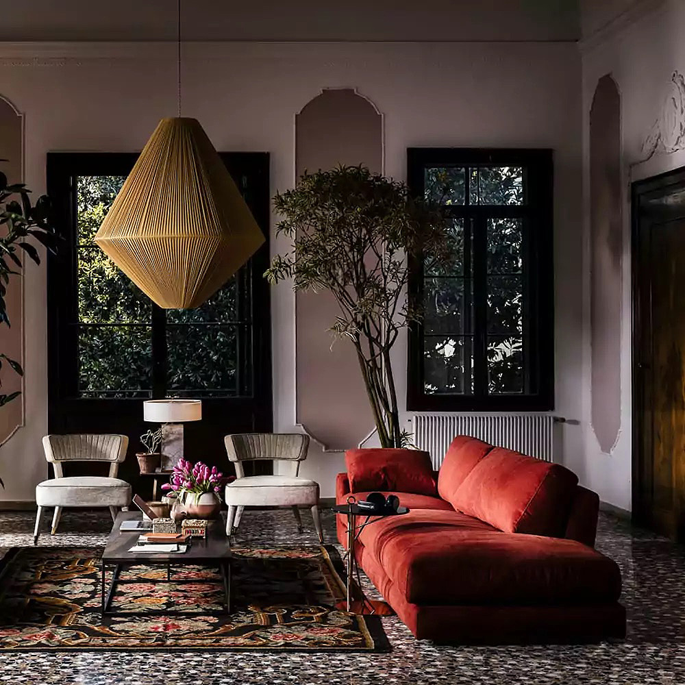 A stylish living room with a red sectional sofa, two white chairs, a black coffee table, and a large window with dark frames. A striking geometric pendant light hangs from the ceiling, and a lush rug with a floral pattern covers the tiled floor. Plants add greenery for those blue Monday moments.