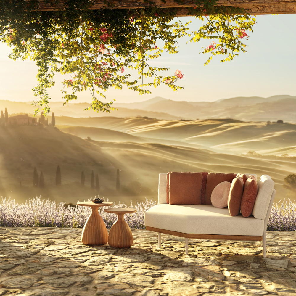 A serene outdoor seating area with a modern beige couch adorned with rust-colored and beige pillows, set on a stone patio overlooking a picturesque landscape of rolling hills at sunset. This luxury exterior features two wooden side tables holding small plants, with vines cascading from above.