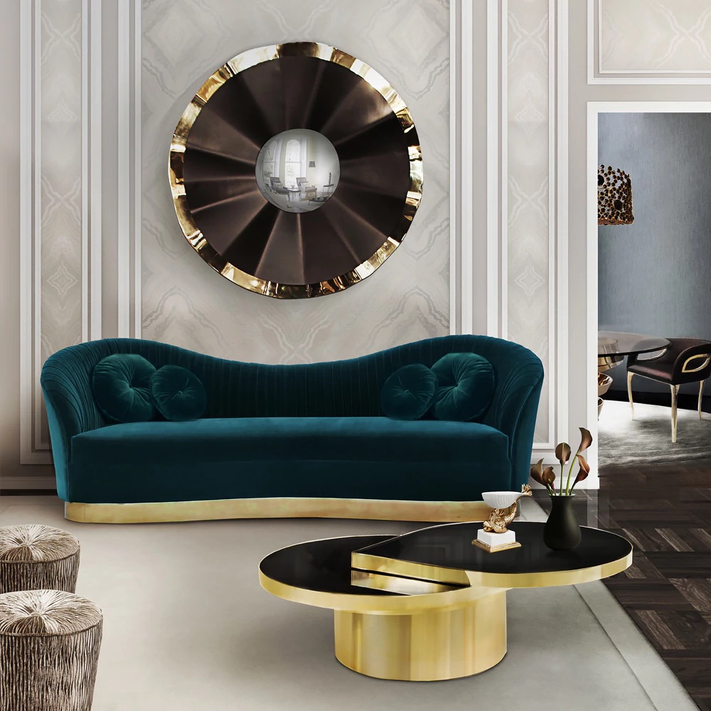 A modern living room featuring a teal, velvet sofa with gold accents and round pillows. A large, circular mirror with a metallic frame hangs on the wall above the sofa. In front of the sofa is a sleek, black and gold coffee table adorned with decorative items.