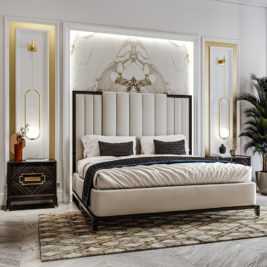 A modern bedroom features a plush, beige upholstered bed with a tall, vertical-panel headboard. Two dark wood bedside tables with geometric details and black and gold decorative lamps flank the bed. The wall behind has a luxurious marble accent, and a patterned rug adorns the floor.