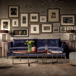 A stylish living room exudes elegance with a dark blue velvet sofa, perfect for easing those "blue Monday" blues. In front, a pair of nested coffee tables holds a vase with yellow tulips, a pink container, a decanter, and glasses. The wall behind the sofa showcases an eclectic collection of framed artwork.